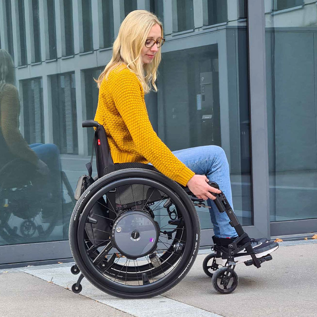 Frau in blauer Jeans und gelbem Pullover bedient Alber e-motion DuoDrive Rollstuhl Fernbedienung